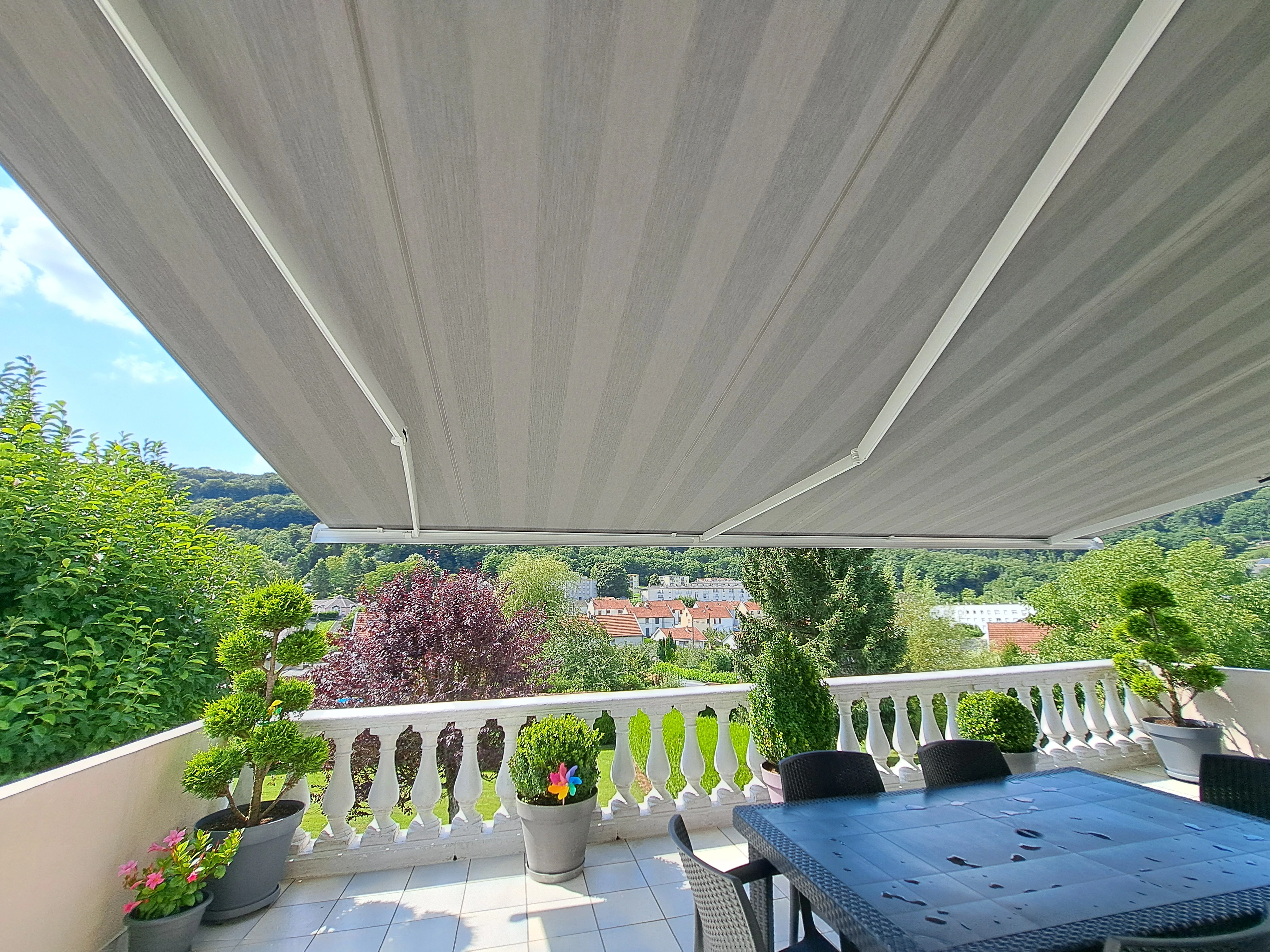 20250805_152143 Vue de dessous d'un store banne gris étendu sur un balcon aménagé avec une table, des plantes et une vue sur la vallée.